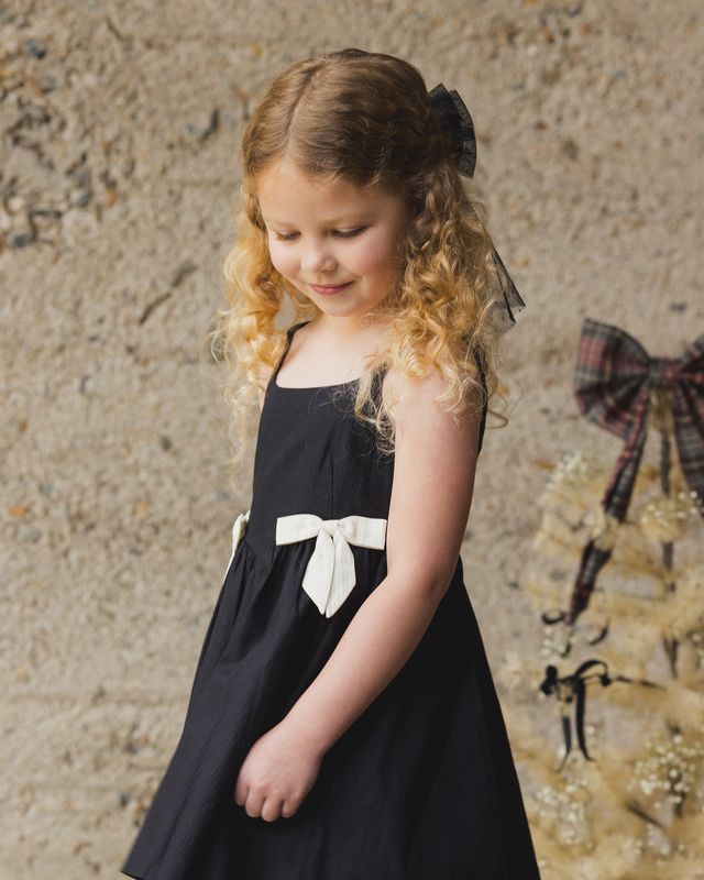 Young girl wearing a black dress with a white bow against a textured wall.