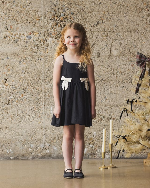 Young girl in a black dress with white bows standing against a textured wall.