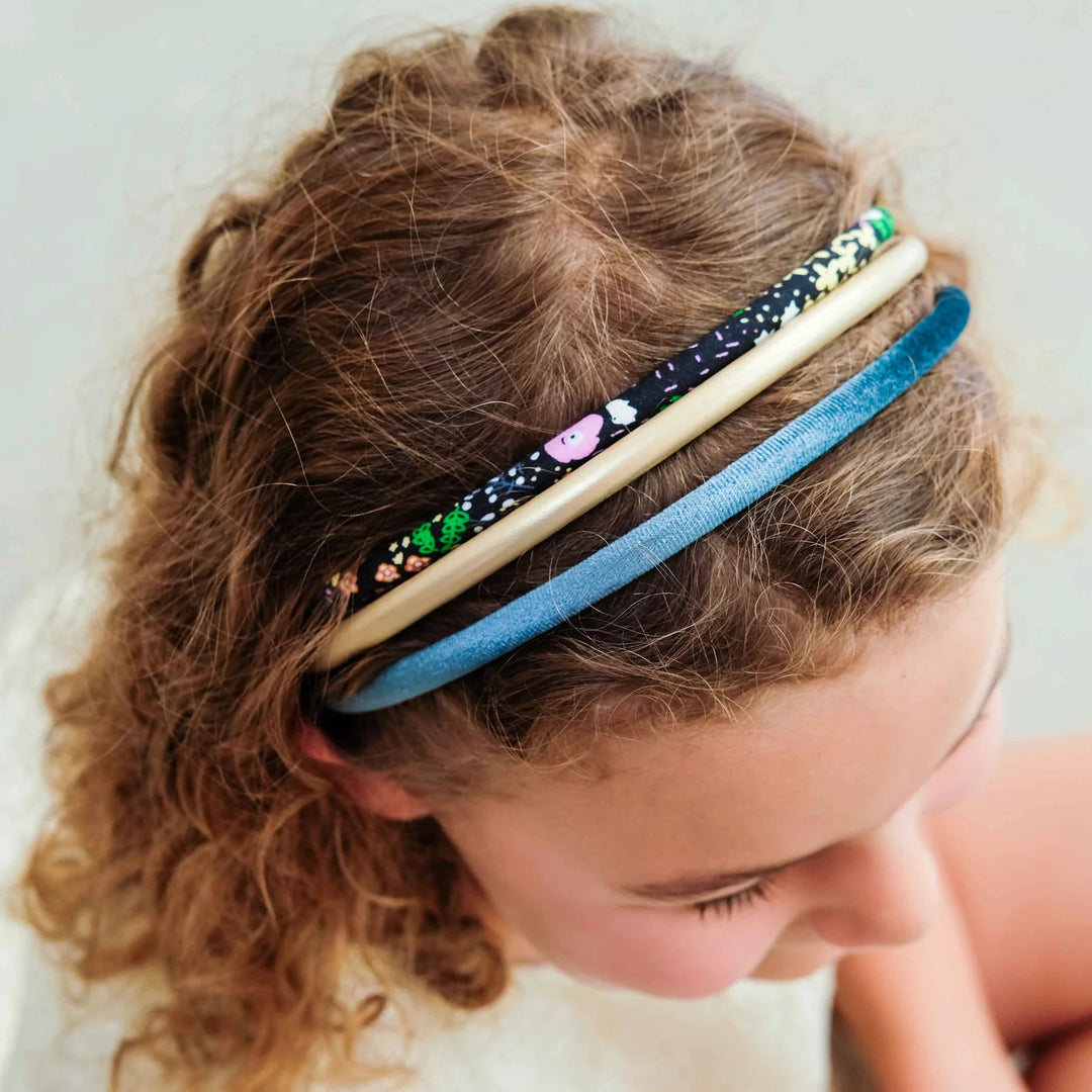 Three fabric headbands in blue, beige, and black with colorful floral and animal prints, worn by a young girl with curly brown hair.