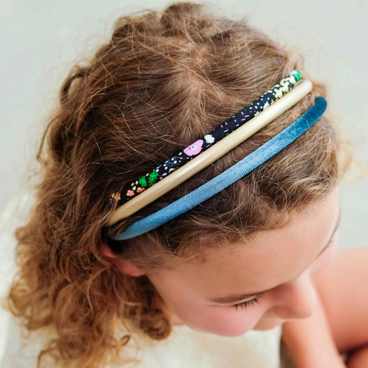 Three fabric headbands in blue, beige, and black with colorful floral and animal prints, worn by a young girl with curly brown hair.