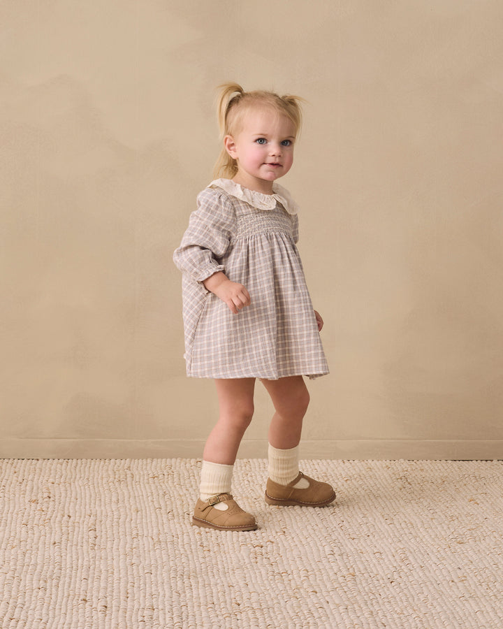 A toddler girl stands in a studio wearing a grey and white gingham dress with a cream collar, cream socks, and tan Mary Jane shoes against a warm beige backdrop.