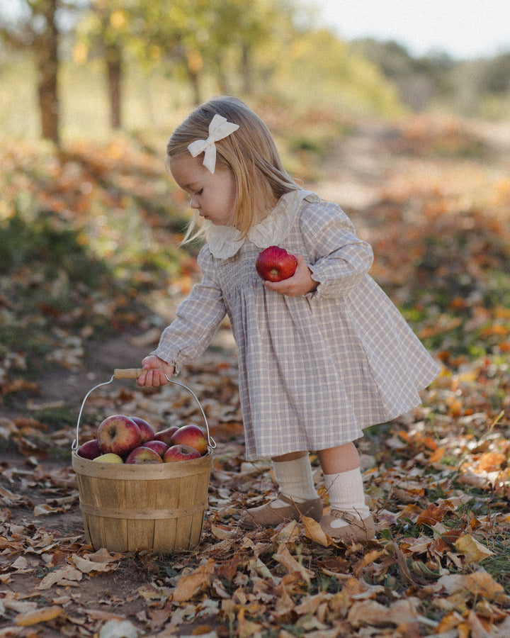 A young girl in a beige and white checkered dress with a white bow in her hair bends down to a wooden basket filled with apples on a leaf-covered path in an autumn park.