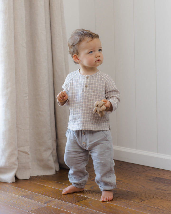 A toddler standing barefoot on a wooden floor indoors, wearing a light gray and white checkered long-sleeve henley top with light gray pants.