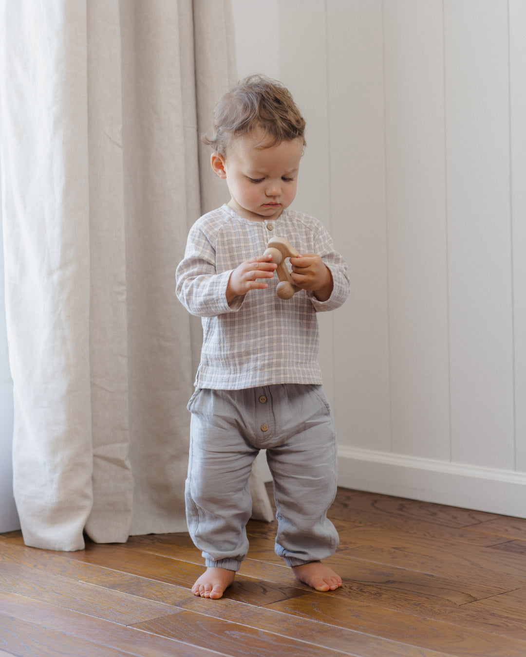 A toddler standing on a wooden floor in a room with white paneled walls and a cream curtain, wearing a gray and white checkered long-sleeve top and light gray pants while holding a small wooden toy.