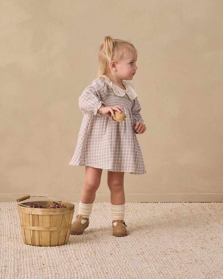 A toddler girl stands in profile wearing a beige and white gingham dress with a white Peter Pan collar, holding a small wooden toy. She is standing on a textured beige rug next to a small wooden basket against a neutral beige background.