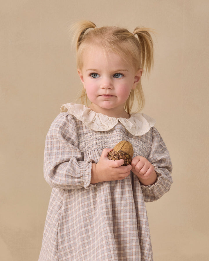 A young blonde girl with pigtails stands against a warm beige studio background, wearing a long-sleeve plaid dress with a cream ruffled collar and holding a small acorn.