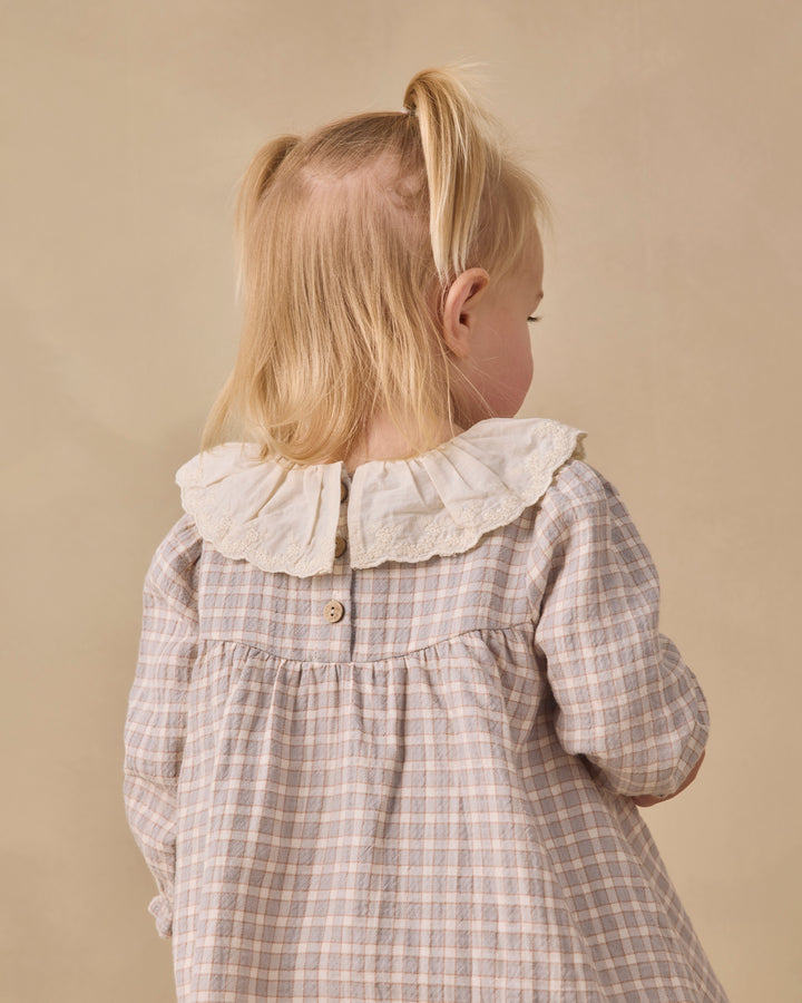 A young blonde girl seen from behind wearing a beige and white plaid dress with a large white ruffled collar, set against a solid beige background.
