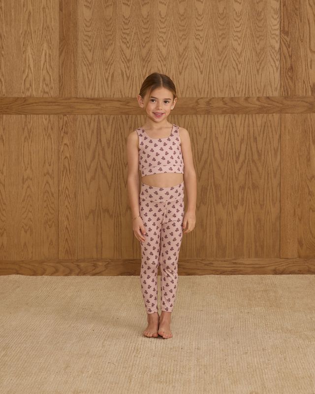 Young girl wearing a matching top and pants set with a floral pattern against a wooden wall.
