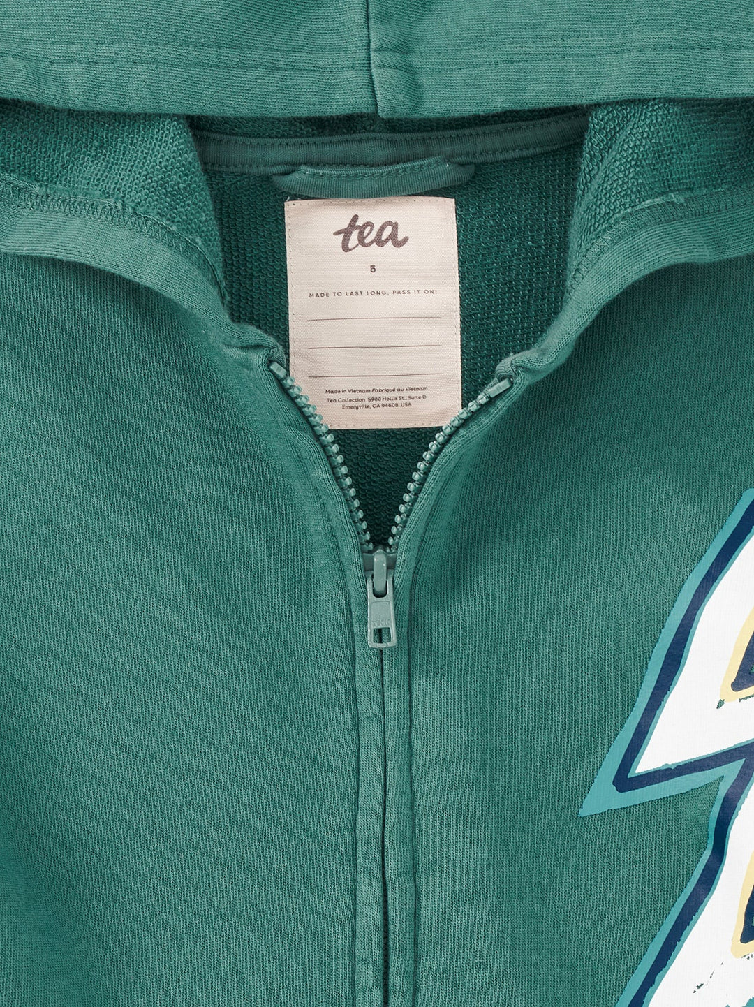 Close-up of a teal green zip-up hoodie showing the inner neck label with 'tea' printed on it, the zipper, and part of a graphic on the right side.