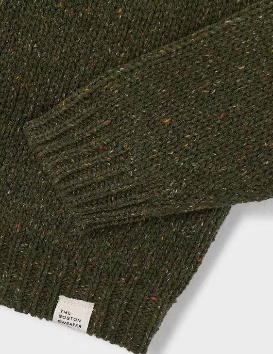 Close-up of an olive green knit sweater with a speckled heather texture and a small white fabric label reading 'THE BOSTON SWEATER' near the cuff.