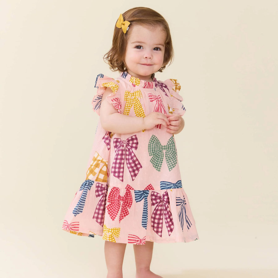 A toddler girl wearing a light pink dress with a colorful bow pattern, standing against a plain beige background.