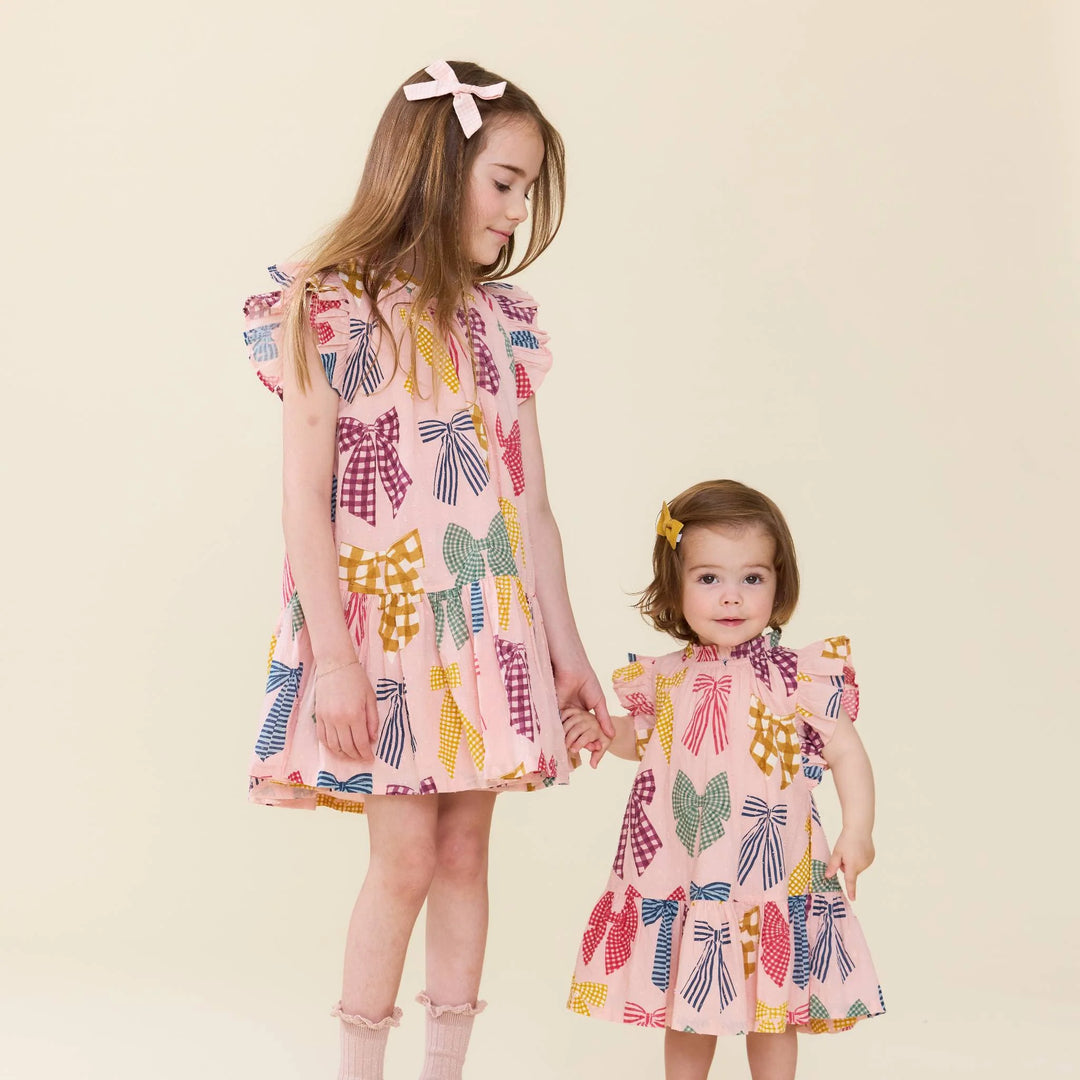 Two young girls wearing matching pink dresses with a colorful bow pattern, standing together against a plain beige background.
