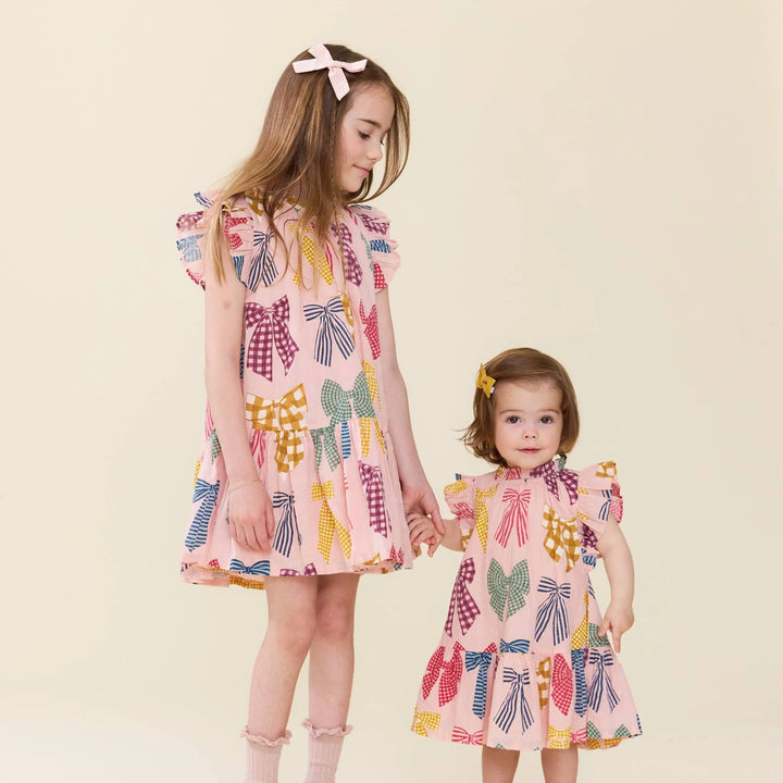 Two young girls wearing matching pink dresses with a colorful bow pattern, standing together against a plain beige background.