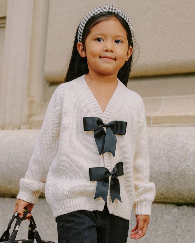 Child wearing a white sweater with black bows in front of a building.