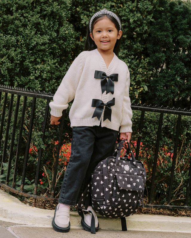 Child wearing a white sweater with black bows, dark jeans, and white shoes, standing next to a polka dot suitcase.