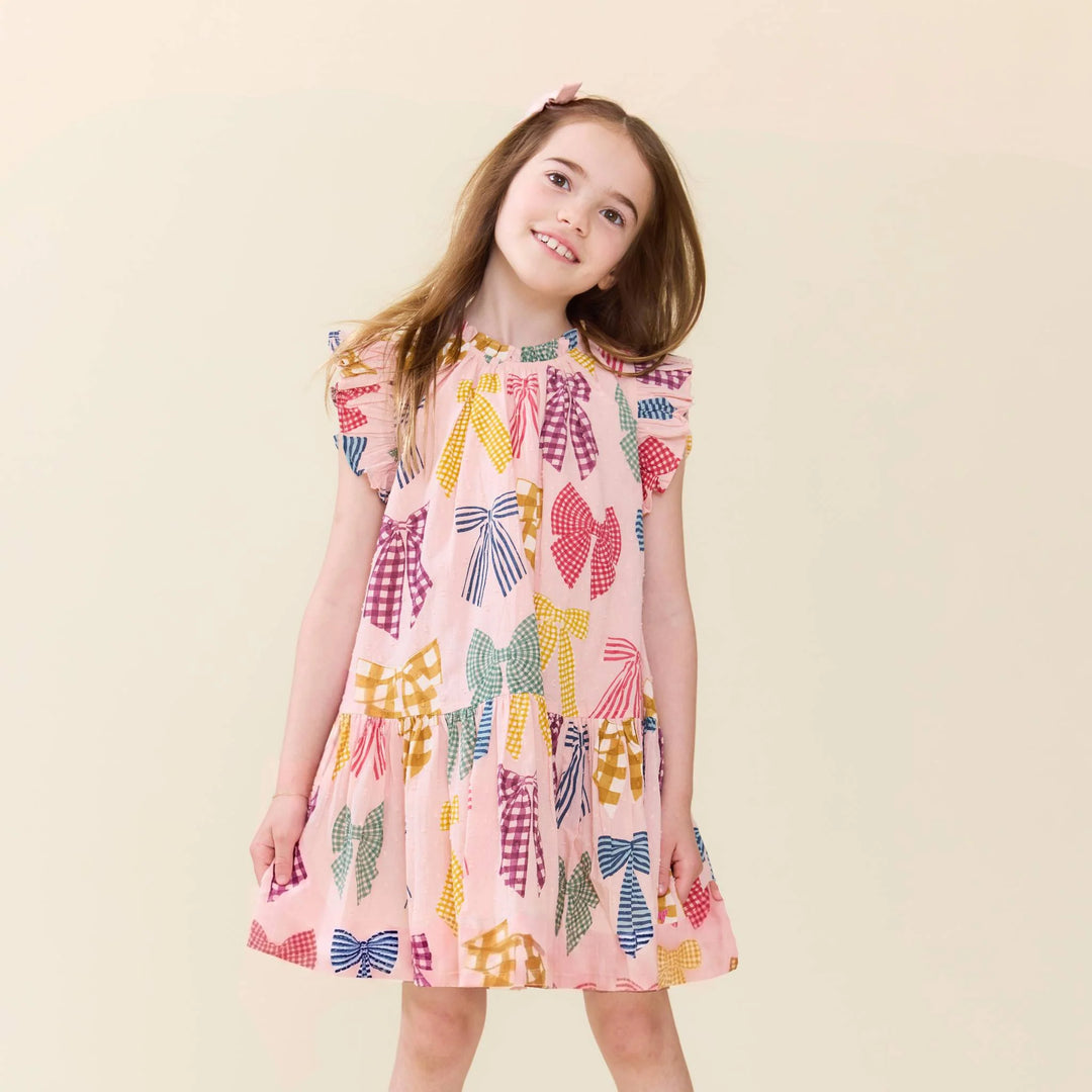 A young girl with long light brown hair wearing a pink dress with a colorful bow pattern, standing against a solid light beige background.