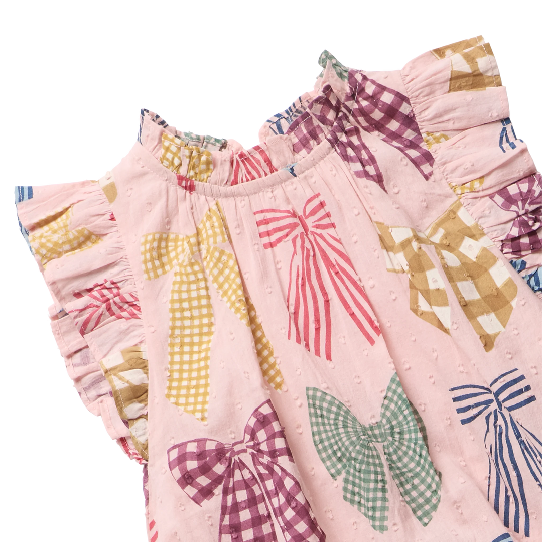 A close-up of a pink dress or tunic with a bow print, featuring ruffled sleeves and waist, decorated with large bow motifs in gingham and striped patterns on a white background.