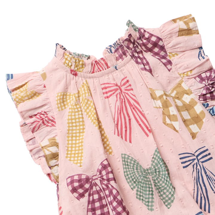 A close-up of a pink dress or tunic with a bow print, featuring ruffled sleeves and waist, decorated with large bow motifs in gingham and striped patterns on a white background.