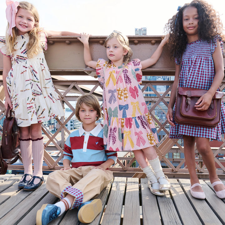Four children modeling coordinated vintage-style clothing and carrying brown leather bags while posing together on a wooden bridge outdoors.