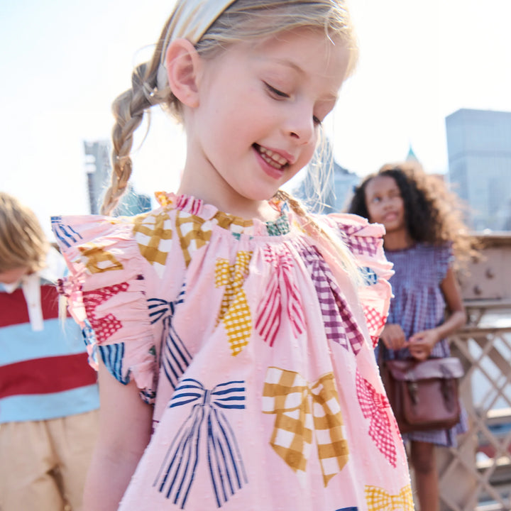 A young girl with a braided ponytail wearing a light pink ruffled blouse with colorful bow and gingham pattern details, photographed outdoors on a sunny day with other children blurred in the background.