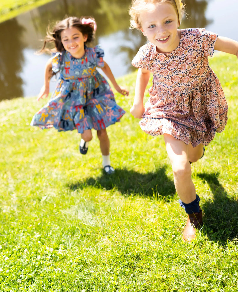 There are two little girls running through the grass. One of the girls is wearing the Jennifer Dress with the bows all over print.  