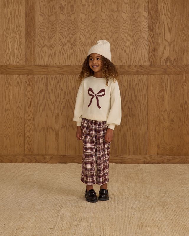 Child wearing a white sweater with a red bow and plaid pants against a wooden paneled wall.