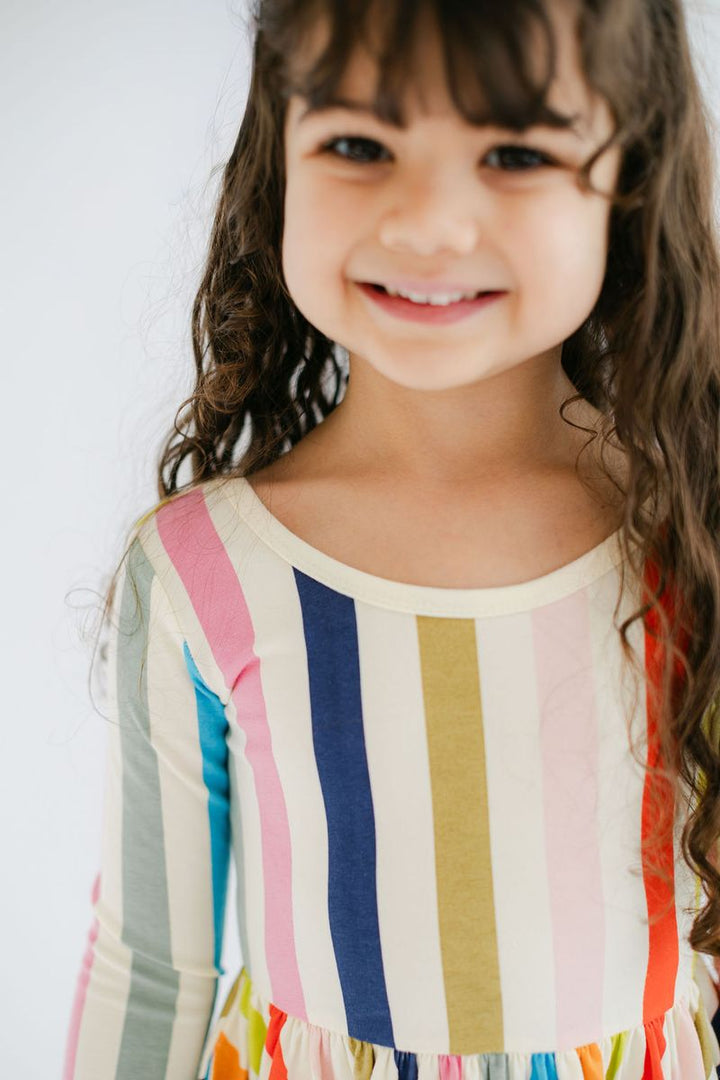 A young girl with long brown hair smiling, wearing a long-sleeve top with vertical stripes in pink, blue, yellow, orange, and green on a cream background.