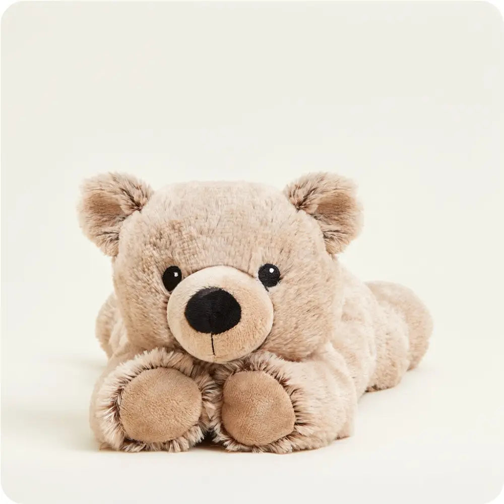 A plush teddy bear toy in a light beige or brown color, lying down and facing forward against a plain, light off-white background.