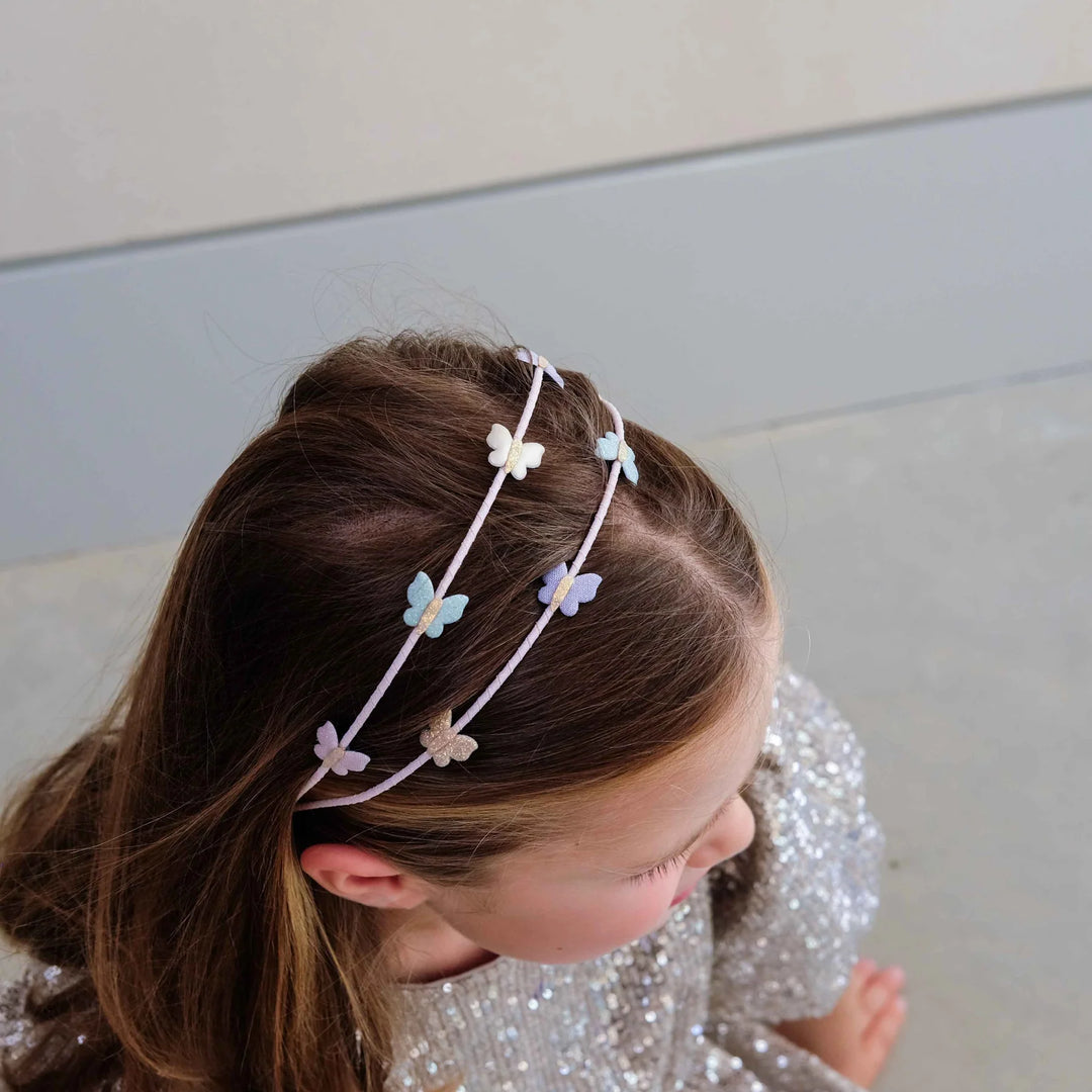 A young girl with brown hair wearing a delicate light purple double-headband adorned with small pastel butterfly charms, paired with a sparkly silver dress.