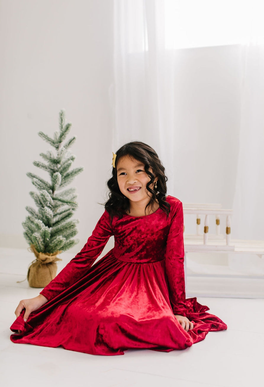 Young girl in a red velvet dress sitting in front of a small Christmas tree.