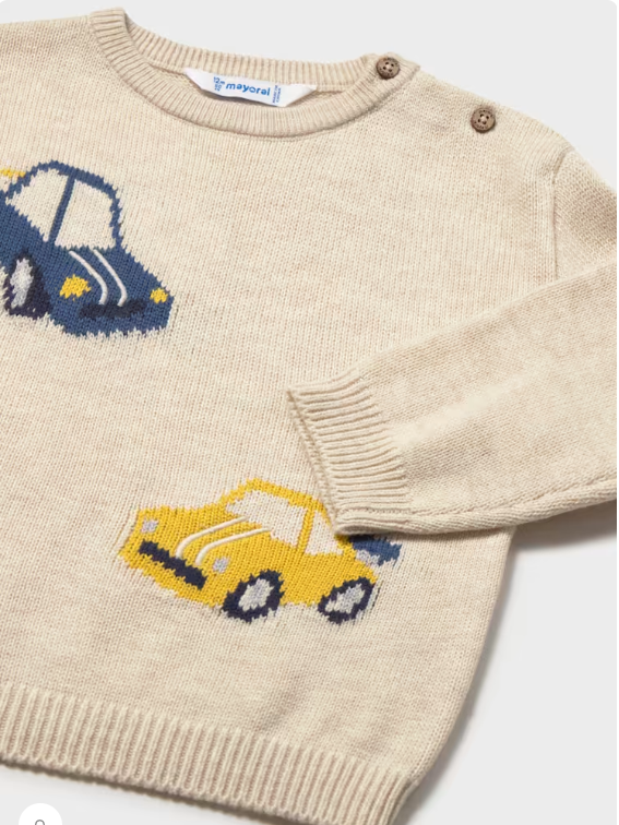 A close-up of a cream-colored knit sweater featuring two knitted cars, one blue and one yellow, on the chest and sleeve. The sweater has a crew neck, ribbed cuffs and hem, and two brown buttons near the shoulder.