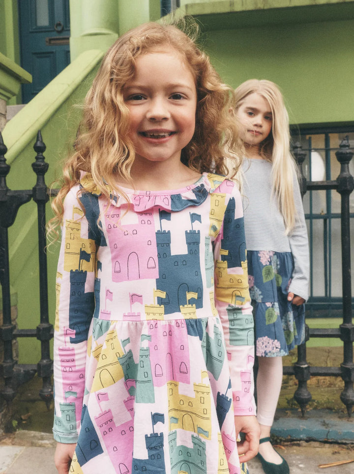 A young girl with curly light brown hair smiles in front of a green house, wearing a long-sleeve dress with a colorful castle pattern in pink, navy, yellow, and blue. Another girl stands behind her.
