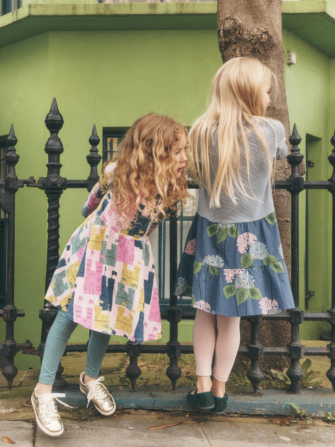 Two young girls standing outdoors in front of a green wall and black metal fence, wearing colorful dresses. One girl has curly light brown hair and wears a pink, yellow, and blue patchwork dress with teal leggings and white sneakers.