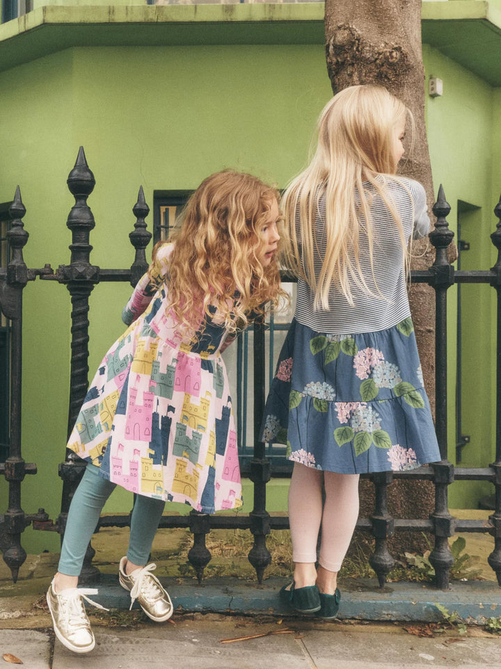Two young girls standing outdoors in front of a green wall and black metal fence, wearing colorful dresses. One girl has curly light brown hair and wears a pink, yellow, and blue patchwork dress with teal leggings and white sneakers.