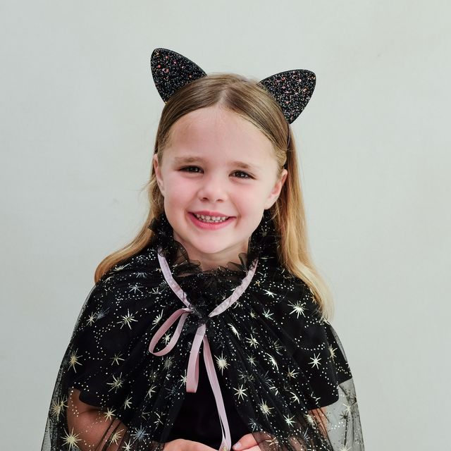 Child wearing a black star-patterned cape and sparkling cat ears on a plain background