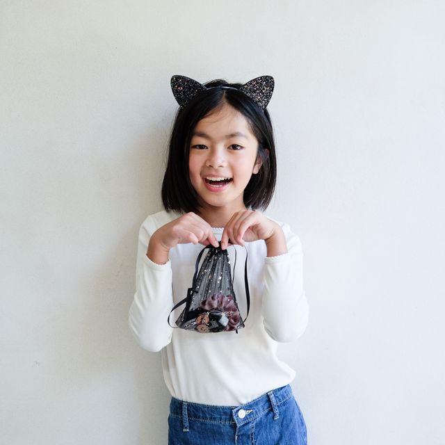 Young girl wearing black cat ears holding a small black bag with a white background