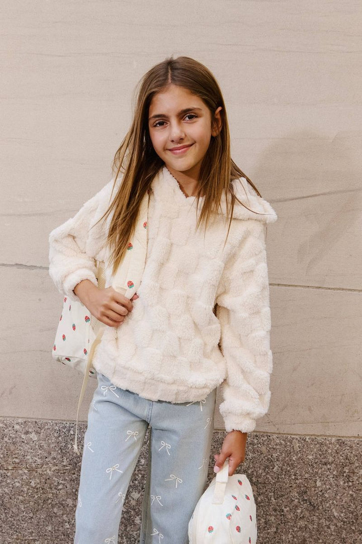Young girl wearing a white textured sweater and light blue jeans, holding a white bag with red polka dots against a neutral wall.