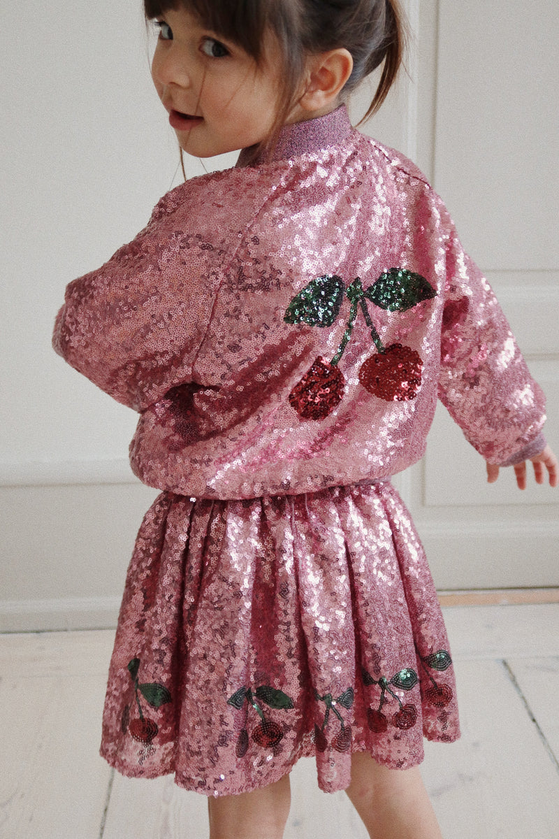 This picture shows the little girl standing from the back with the sequin jacket on. The jacket is pink with large cherries on it. 