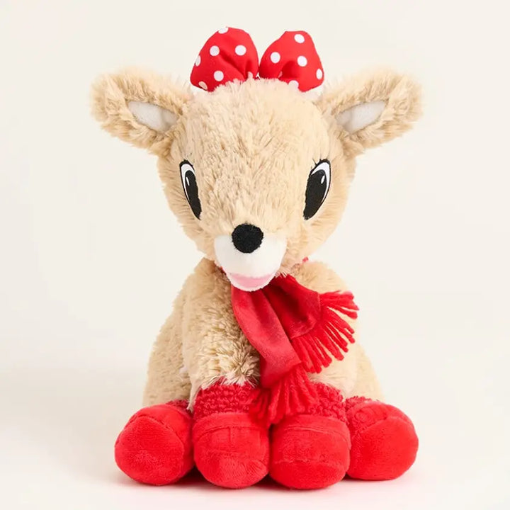 A plush toy deer sitting against a plain light background, with a beige body, large black eyes, a red bow with white polka dots on its head, a red scarf, and red booties.