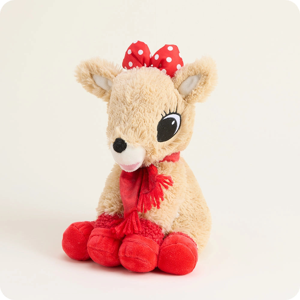 A plush toy deer in light beige faux fur, wearing a red bow with white polka dots and a red scarf, sitting against a plain off-white background.