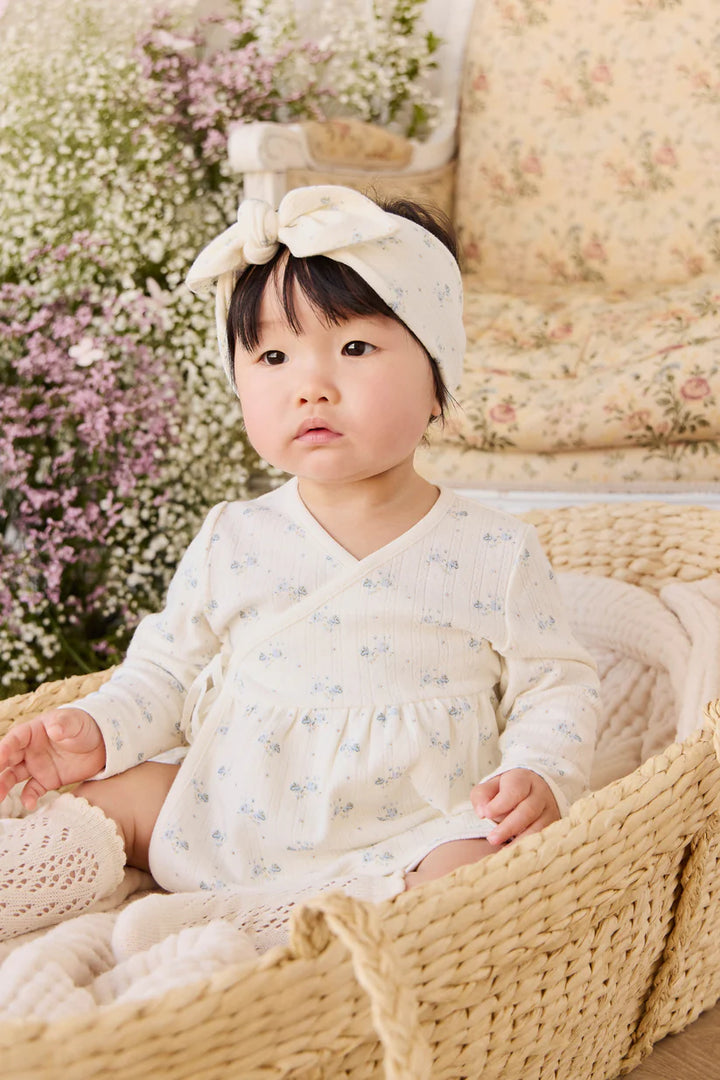 Baby in a white outfit with a bow sitting in a basket surrounded by flowers and a floral-patterned couch.