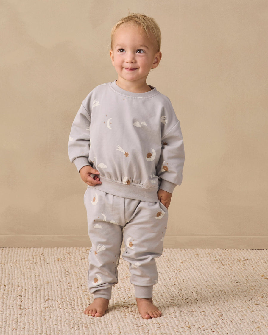 A toddler stands barefoot on a textured cream rug against a warm beige studio wall, wearing a light gray long-sleeve sweatshirt and matching jogger pants with small scattered animal illustrations.