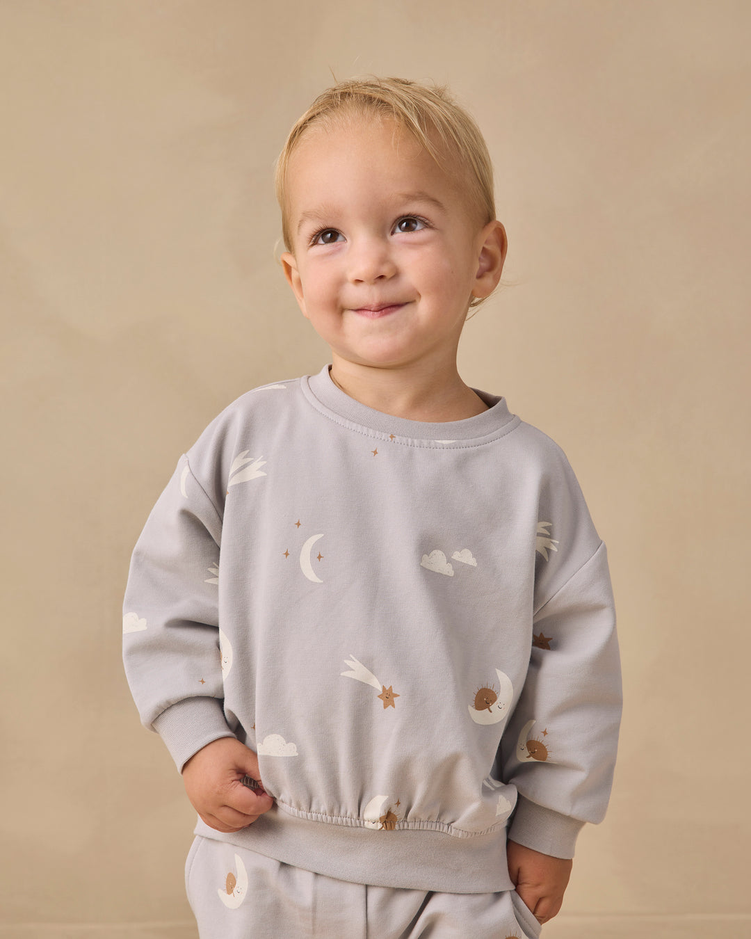 A smiling toddler with blonde hair wearing a light gray long-sleeve sweatshirt and matching pants with a celestial print of white clouds, moons, and stars, set against a plain beige background.