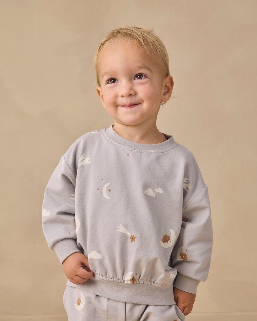 A smiling toddler with blonde hair wearing a light gray long-sleeve sweatshirt and matching pants with a celestial print of white clouds, moons, and stars, set against a plain beige background.