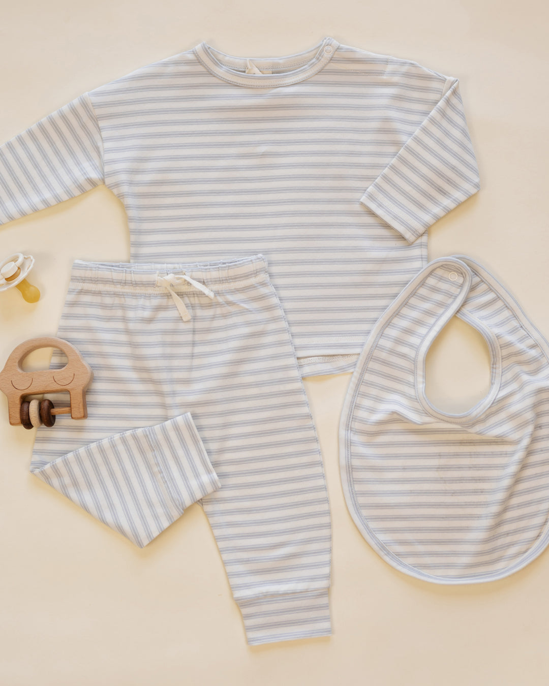 A baby clothing set laid flat on a warm cream background, including a long-sleeve top, matching pants, and a bib, all in a light gray and white stripe pattern. A small wooden pacifier clip is also placed beside the set.