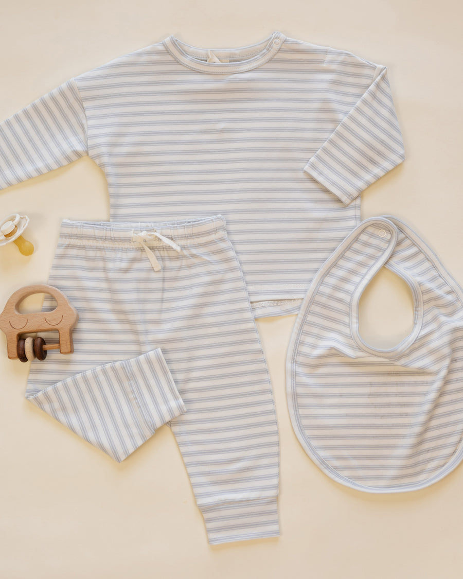 A baby clothing set laid flat on a warm cream background, including a long-sleeve top, matching pants, and a bib, all in a light gray and white stripe pattern. A small wooden pacifier clip is also placed beside the set.