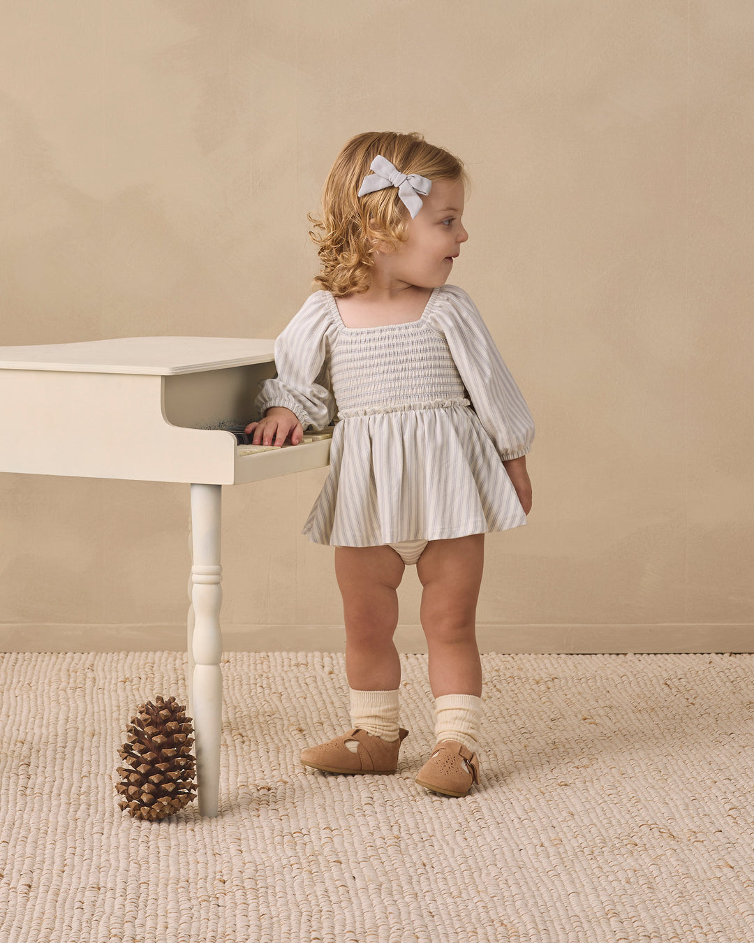 A toddler girl stands beside a small white table in a neutral studio setting. She is wearing a smocked long-sleeve dress with vertical white and gray stripes, cream socks, tan shoes, and a white bow in her hair.