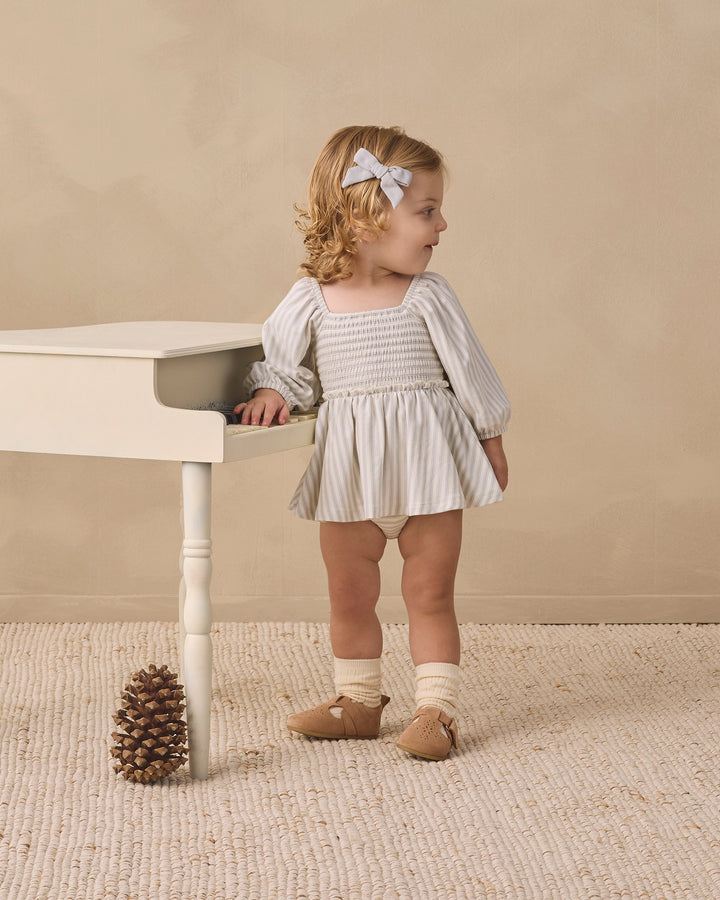 A toddler girl stands beside a small white table in a neutral studio setting. She is wearing a smocked long-sleeve dress with vertical white and gray stripes, cream socks, tan shoes, and a white bow in her hair.