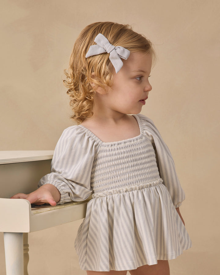 A young girl with blonde curly hair wearing a cream and white vertically striped smocked dress with puffed sleeves and a matching bow in her hair, posed next to a white piano against a beige background.