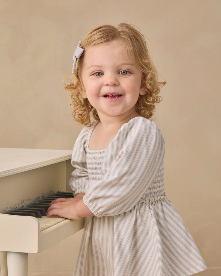 A young girl with curly blonde hair wearing a cream and gray striped dress stands next to a white piano against a warm beige background.